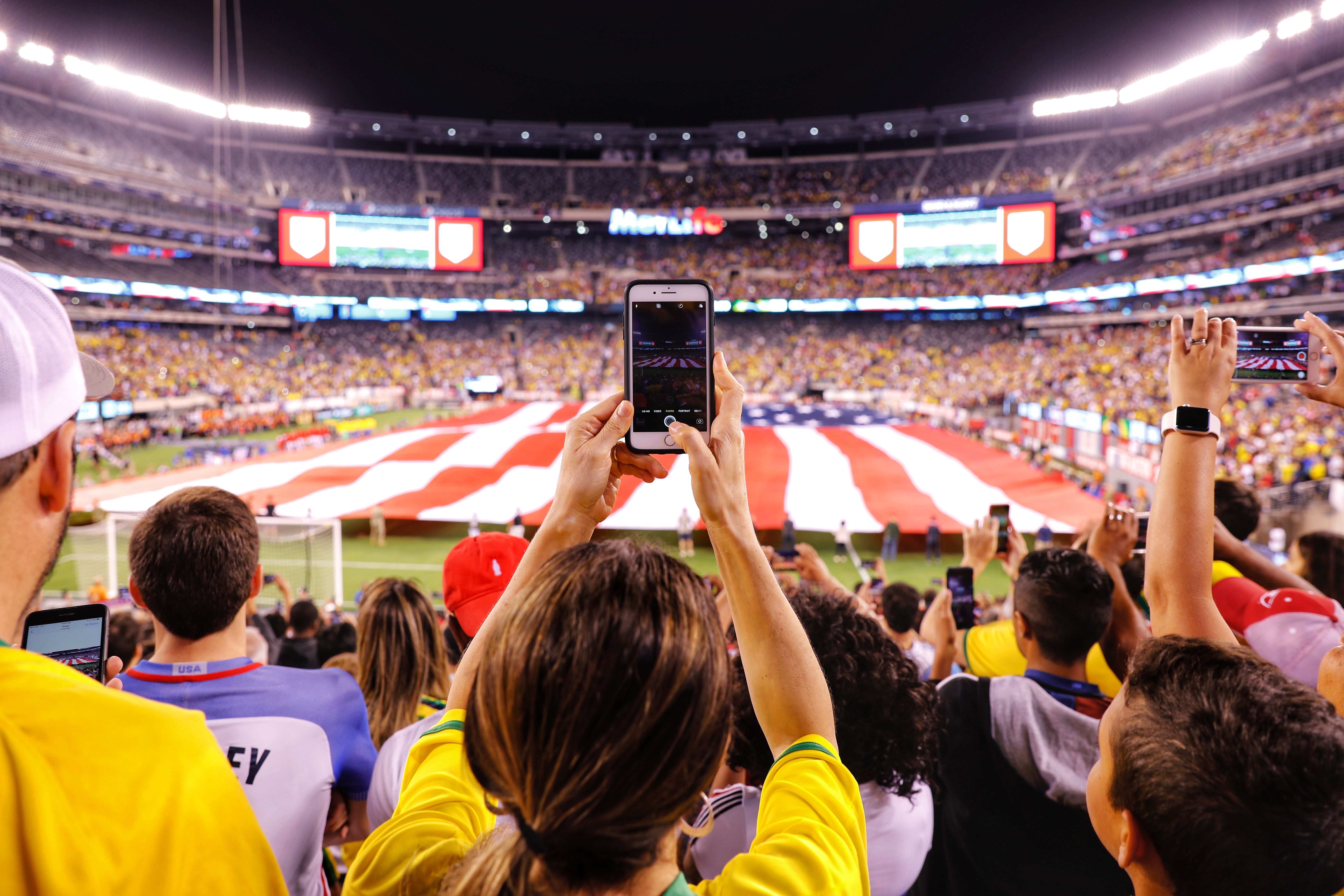 
		People in a stadium at a football game holding up their cell phones to record a huge American flag presentation at Halftime.